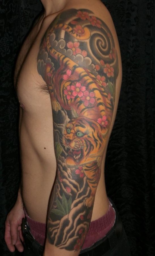 Tatuaje de estilo manga, es decir, que cubre el brazo al completo con la image de un tigre adornado de coloridas flores rosas y un fondo de nubes negras